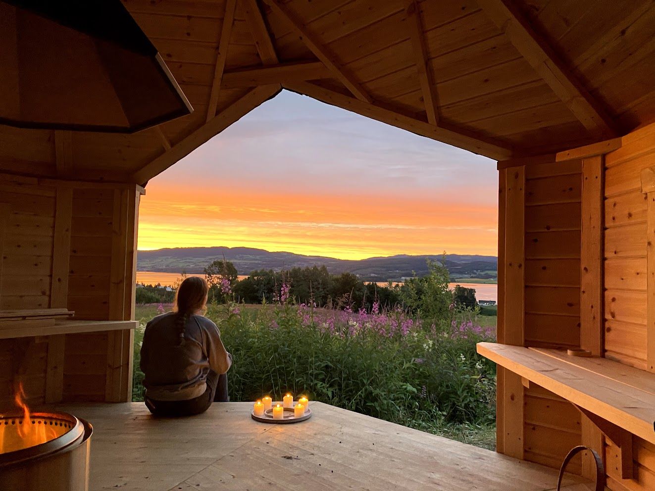 Utsikt fra gapahuken på gården mot Mjøsa i solnedgang, med bålpanne og telys